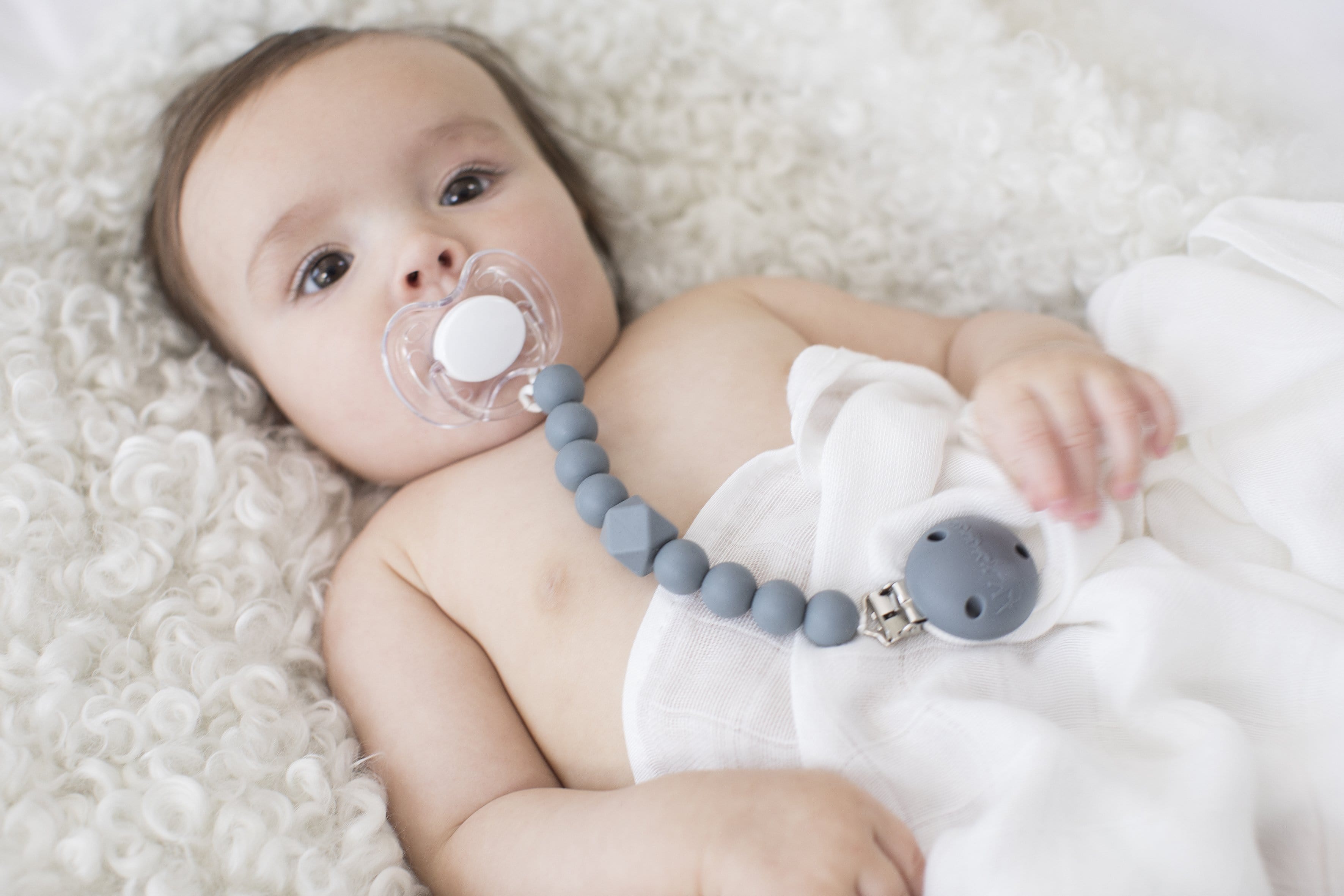 The Les Enfants Chewy Pacifier Clip grey with a baby model and white les enfants 100% bamboo muslin blanket