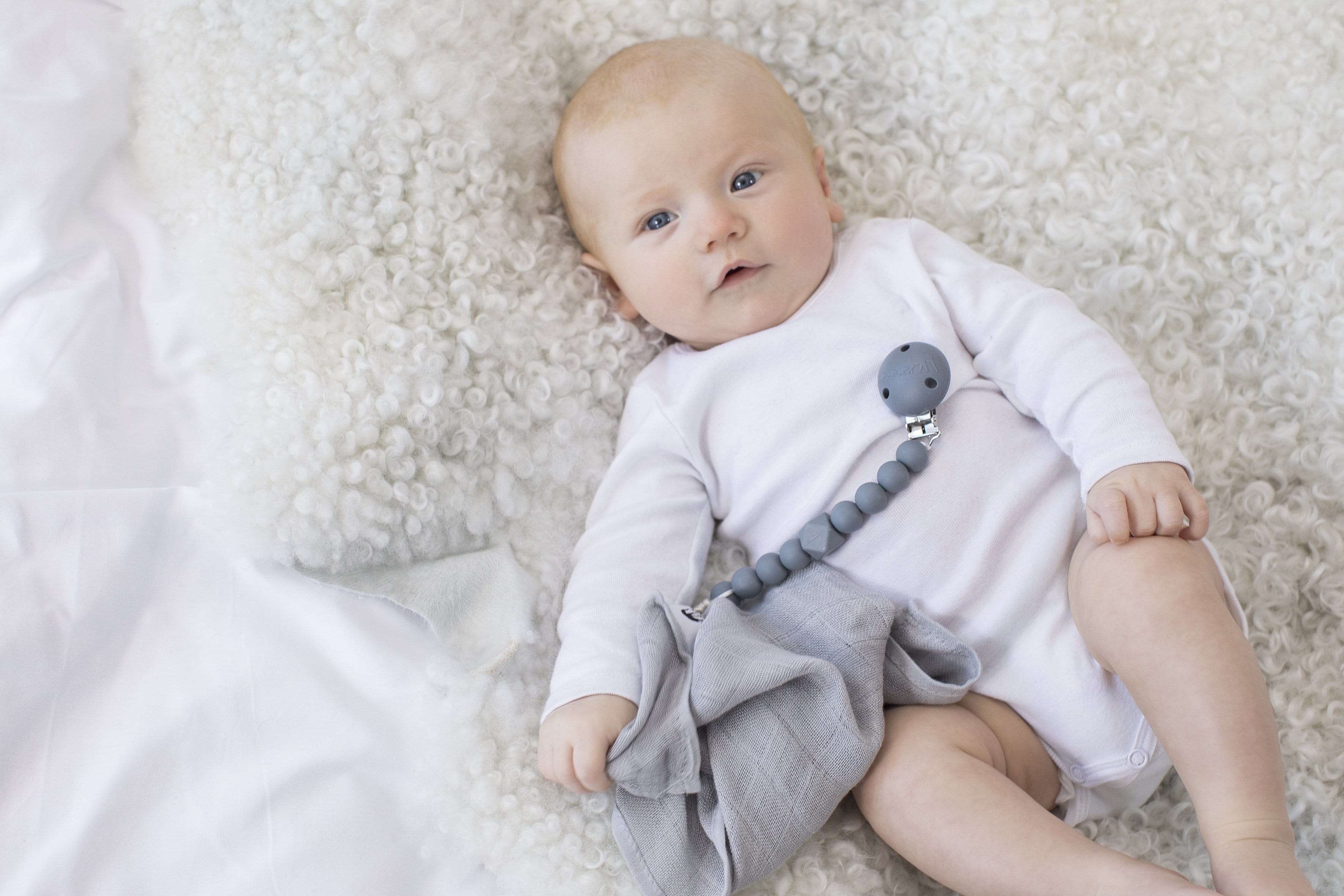 The Les Enfants Chewy Pacifier Clip grey with a blond baby model and grey les enfants 100% bamboo muslinblanket